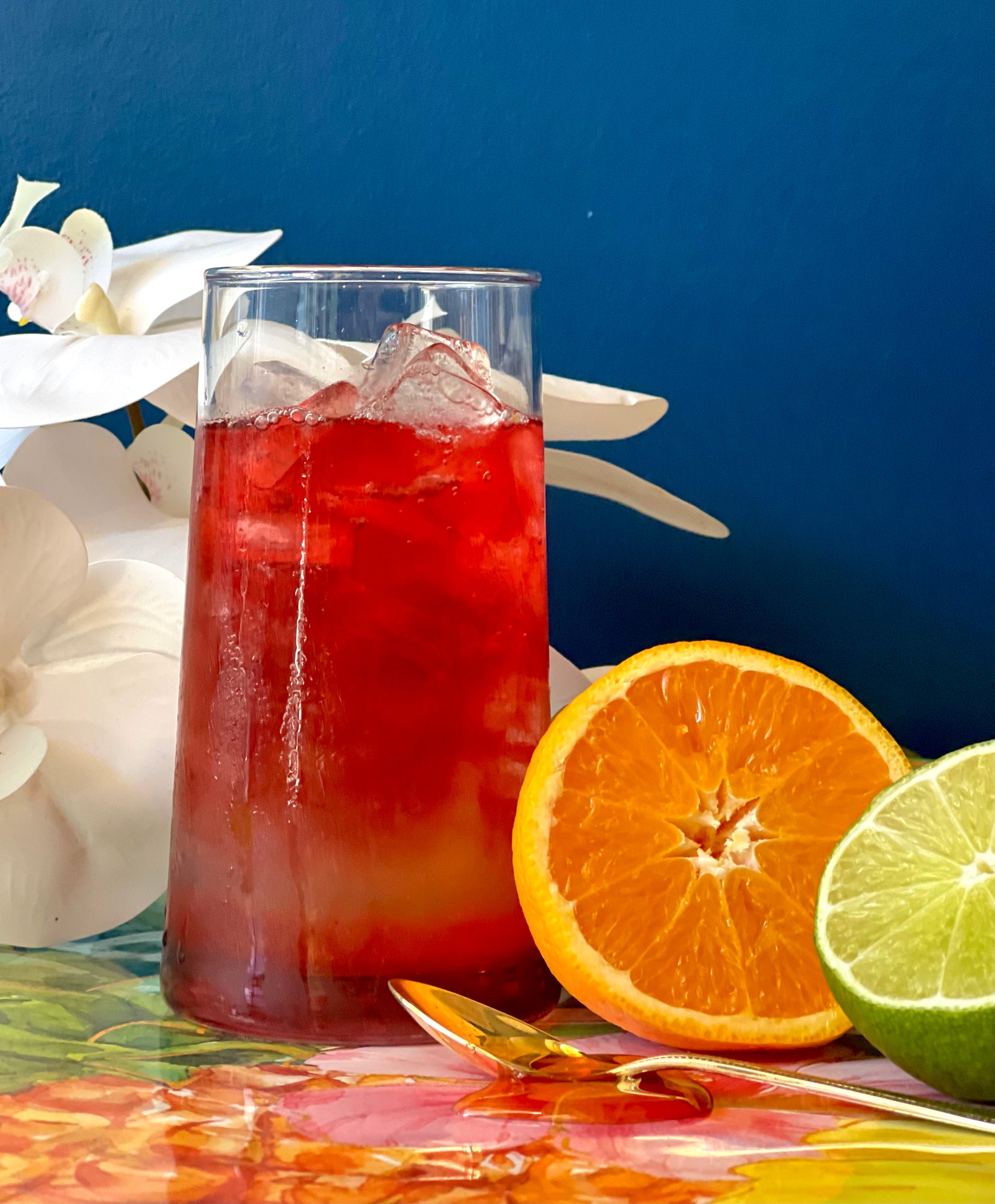Red cocktail in a glass with ice, surrounded by sliced oranges and limes on a reflective surface.