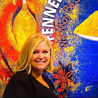 A smiling blonde woman stands in front of a colorful wall mural for 'Flannel & Fire Crafting House,' featuring a large spoonful of yellow powder and bold white and orange text on a blue background.