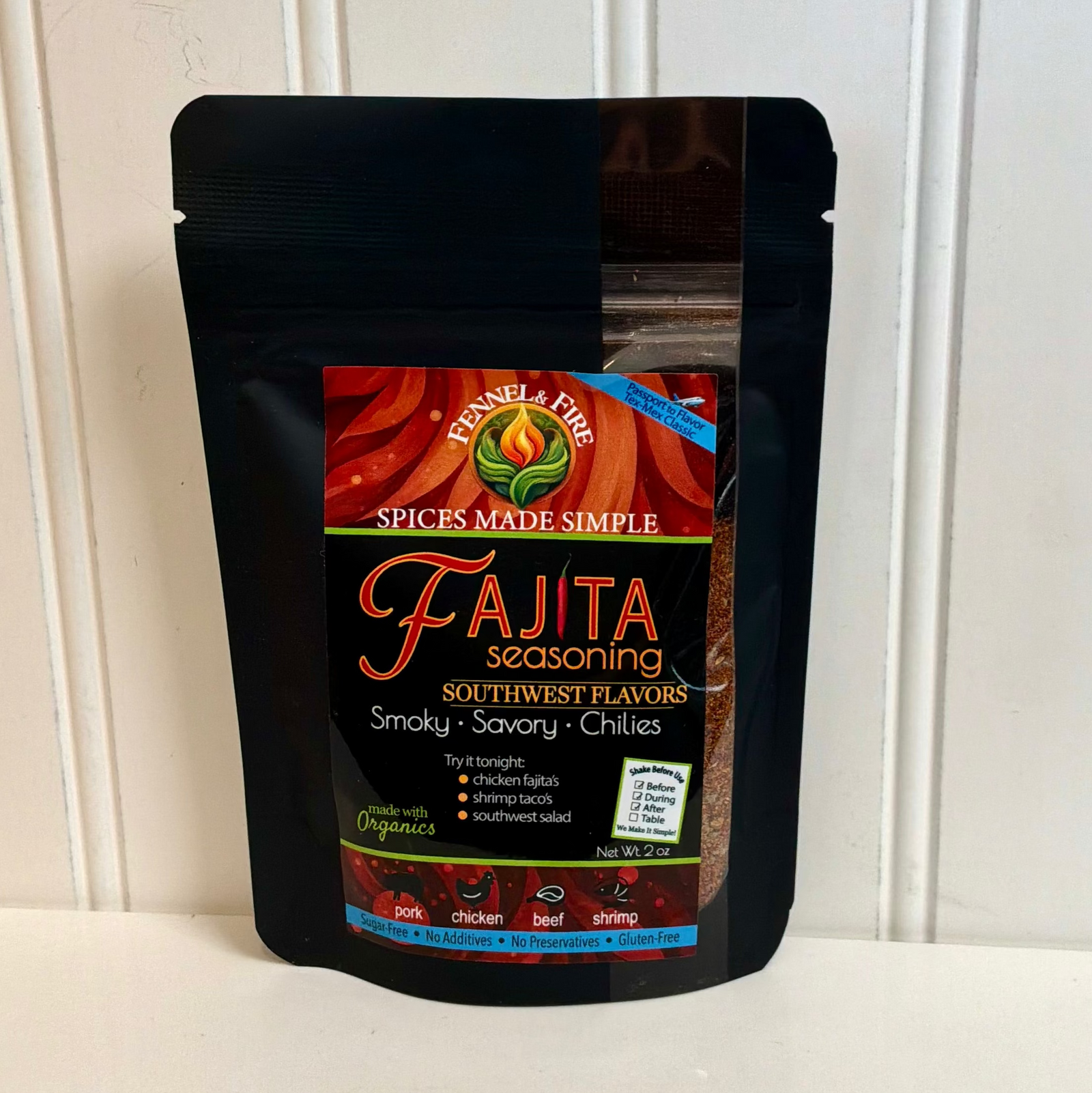 A black resealable stand-up pouch of Fajita Seasoning by Spice & Fire, standing on a white surface against a white paneled wall.
