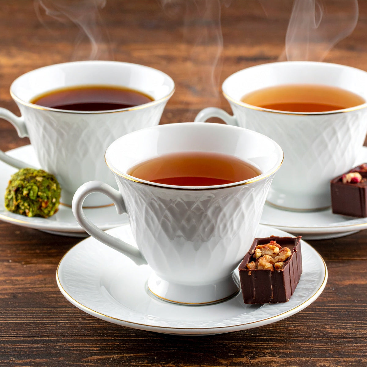 Three white teacups filled with steaming tea on a wooden table with saucers and small desserts.