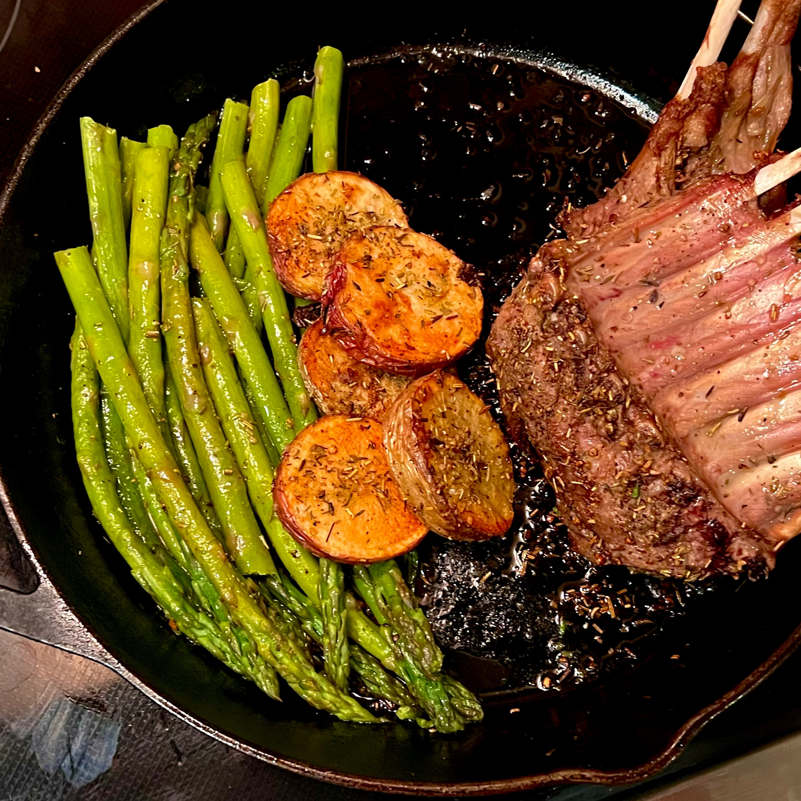 Lamb rack with asparagus and potatoes in a cast iron skillet on a stove.