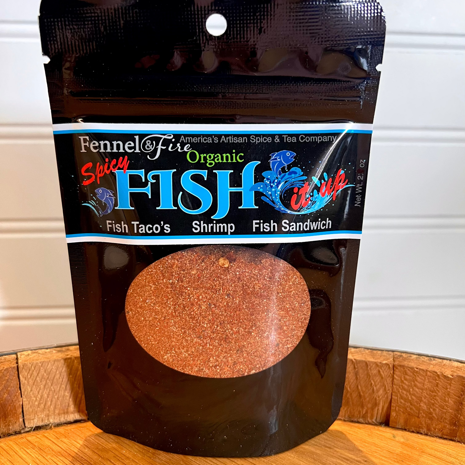 Fennel & Fire Organic Shitty Fish it up spice package on a wooden surface with a white wall background.