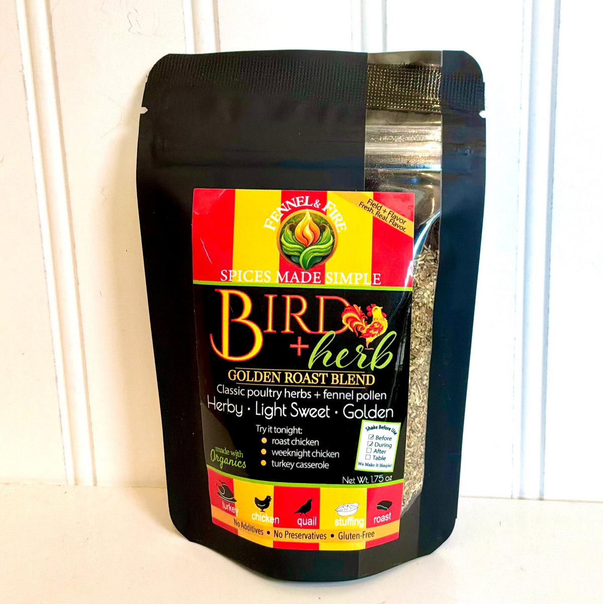 A black stand-up pouch of Fancy A Fire Bird + Herb Golden Roast Blend spice mix, standing on a white surface against a white paneled wall.