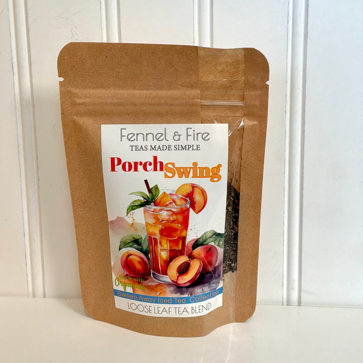 A brown kraft paper stand-up pouch of Fennel & Fire Teas Made Simple 'Porch Swing' loose leaf tea blend, displayed on a white surface against a white paneled wall.