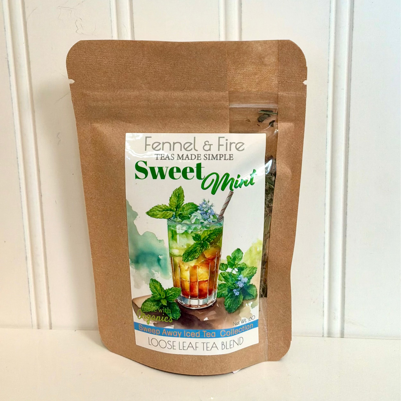 A brown kraft paper stand-up pouch of Fennel & Fire Sweet Mini loose leaf tea, featuring a colorful label with a glass of iced tea and mint leaves, set against a white paneled background.